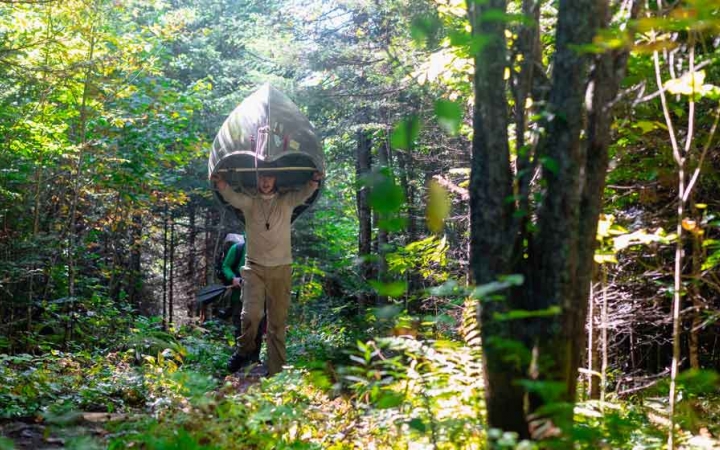 a student portages a canoe along a wooded trail on an outward bound course